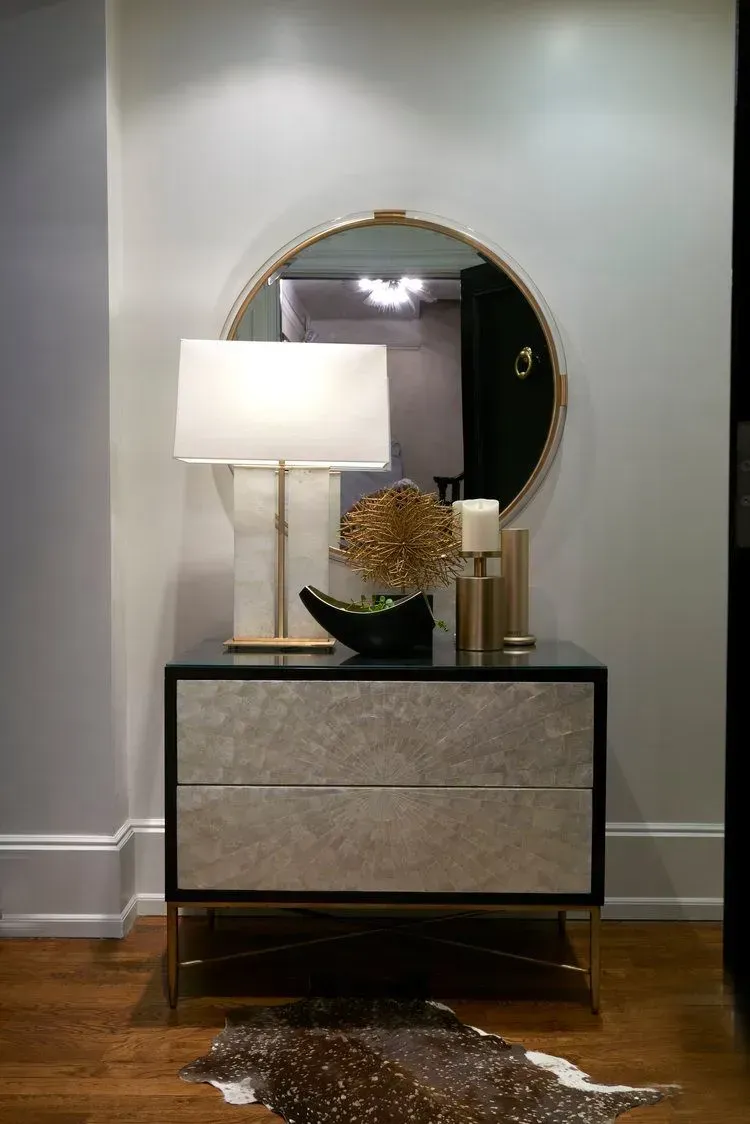 Nightstand with lamp, mirror, and decor. Silver drawers on black frame, gold accents, cowhide rug.