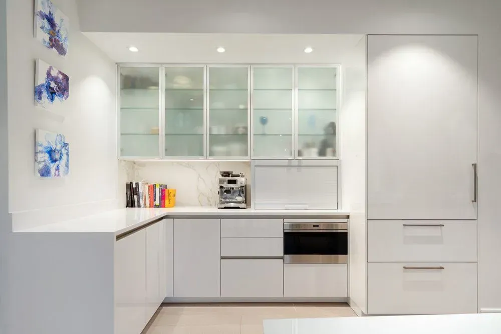 Modern white kitchen with glass-front cabinets, built-in oven, and a refrigerator. Artwork hangs on the wall.