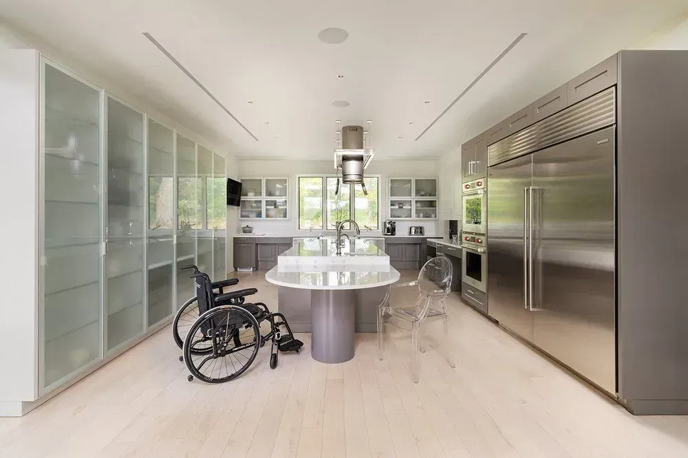 Wheelchair in modern accessible kitchen with white cabinetry and large stainless steel refrigerator.