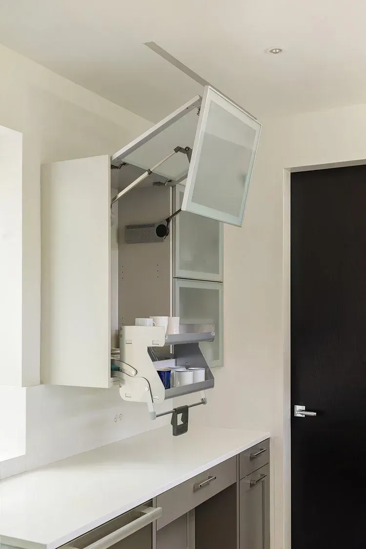 White cabinet with upward-opening glass doors in a kitchen. Open cabinet reveals medication organizers.