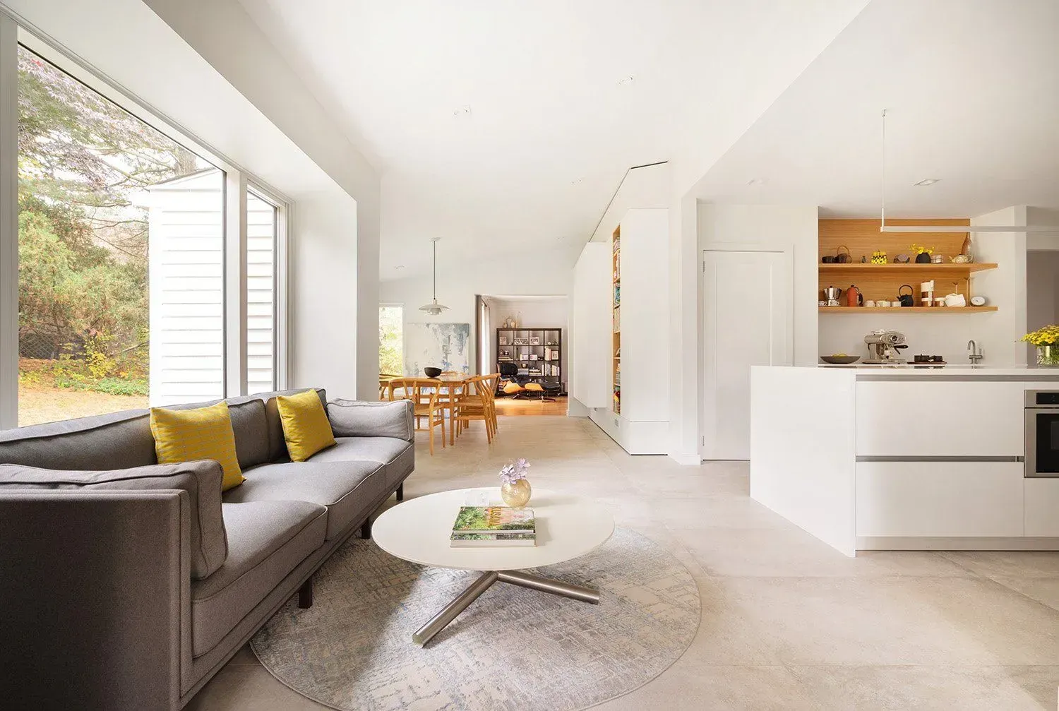 Bright, modern living space with gray sofa and yellow pillows, round coffee table, open to dining area and kitchen.