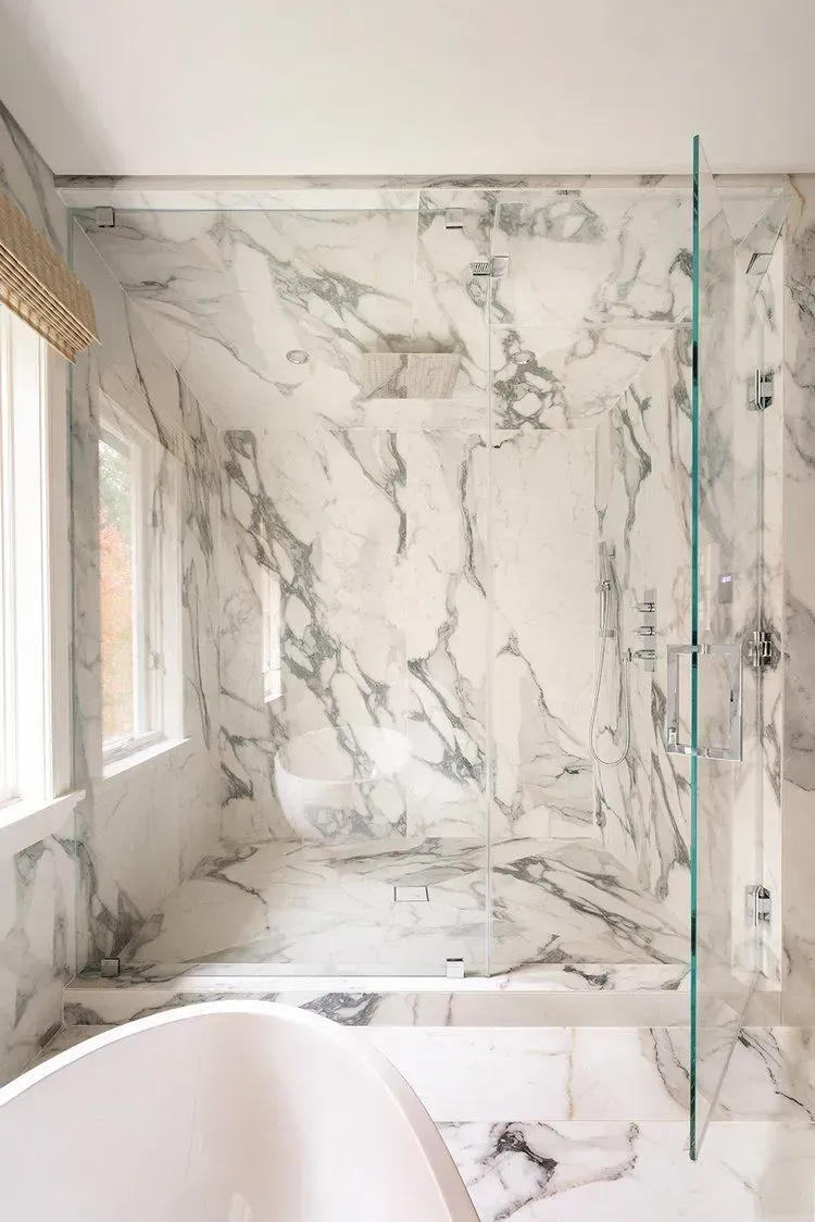 Marble-tiled shower with glass door in a bathroom. A window with a blind is to the left, and a tub is in the foreground.