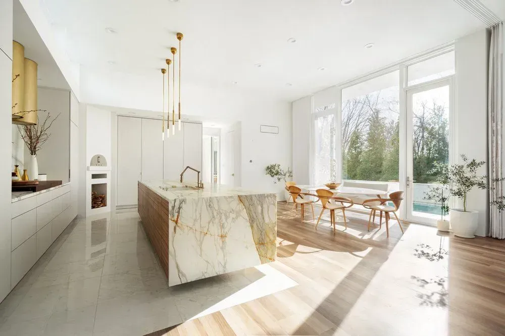 Bright, modern kitchen with large island, marble countertop, and wood cabinets. Dining area with natural light and outdoor view.