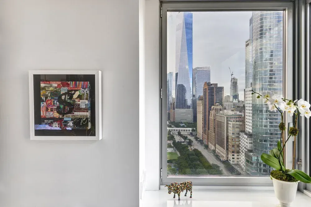 Artwork on wall next to a window overlooking a cityscape. White flowers and small figurine on windowsill.