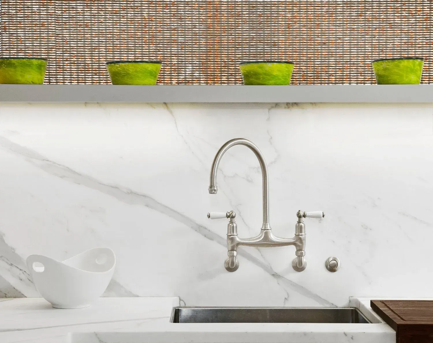 Kitchen sink with marble backsplash, silver faucet, and green bowls on a shelf.