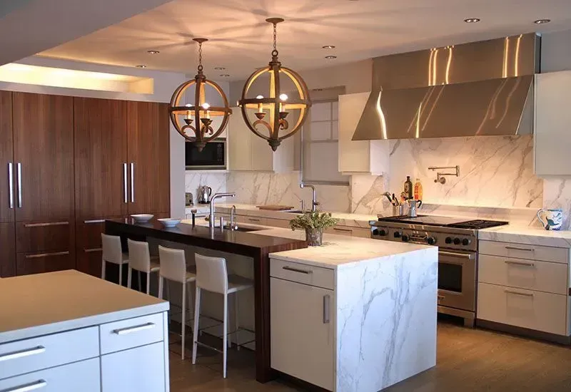 Modern kitchen with marble countertops, stainless steel appliances, and wood cabinetry. Two spherical pendant lights hang above an island with bar seating.