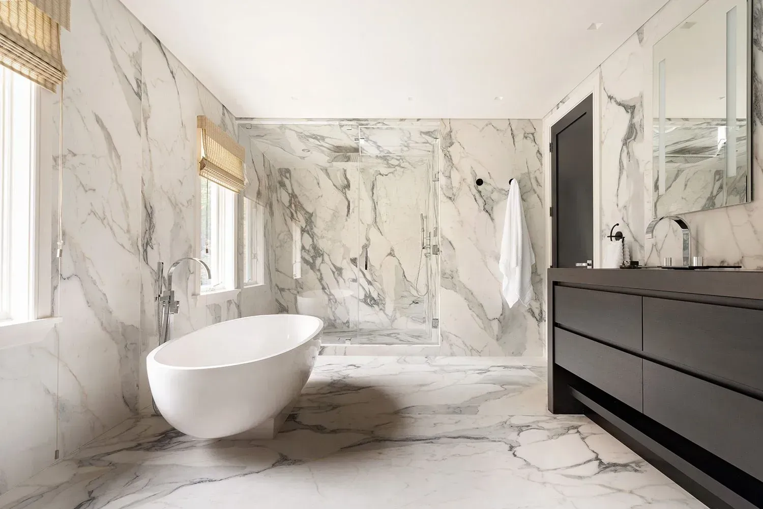 Luxurious white marble bathroom with standalone tub, shower, dark vanity, and window.