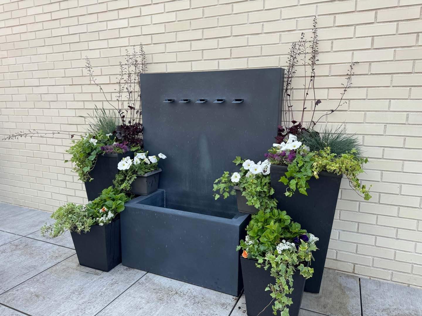 A fountain is surrounded by potted plants in front of a brick wall.