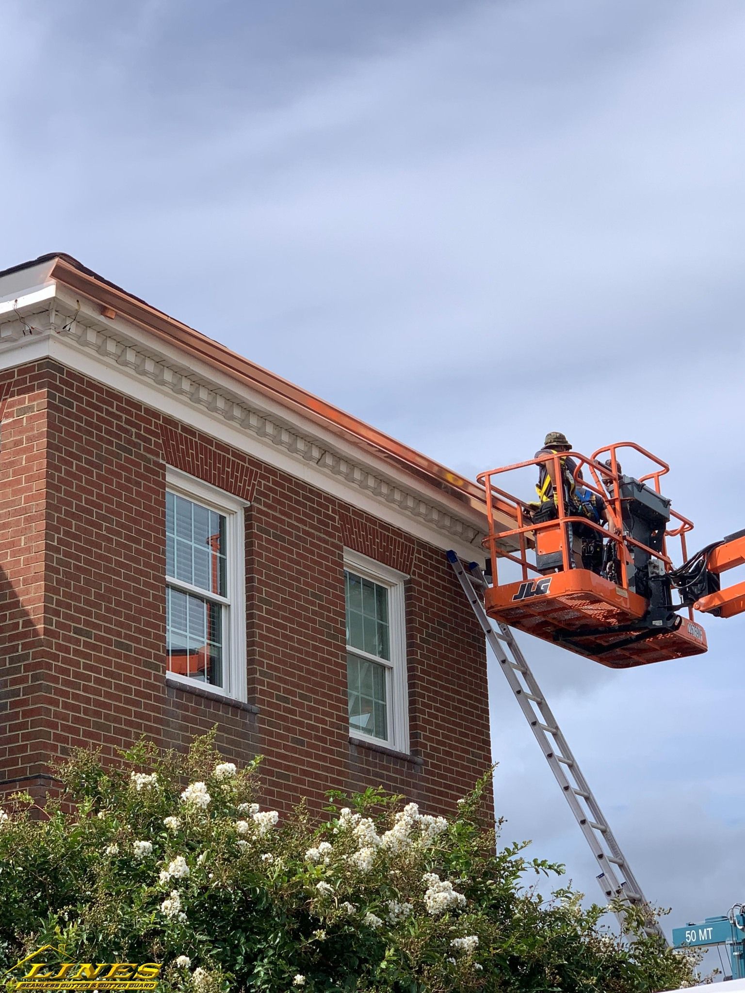 Person in an orange lift working on the roof of a brick building with white-trimmed windows.