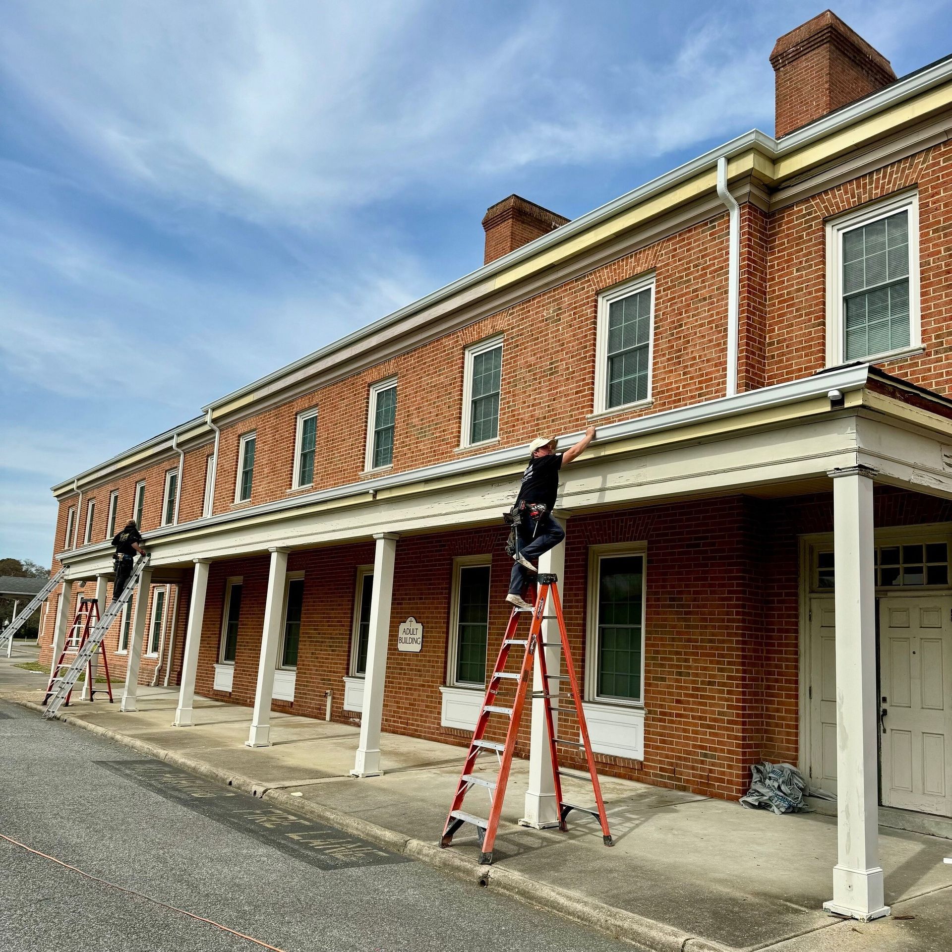 Two people on ladders cleaning gutters on a brick building with white columns under a blue sky.