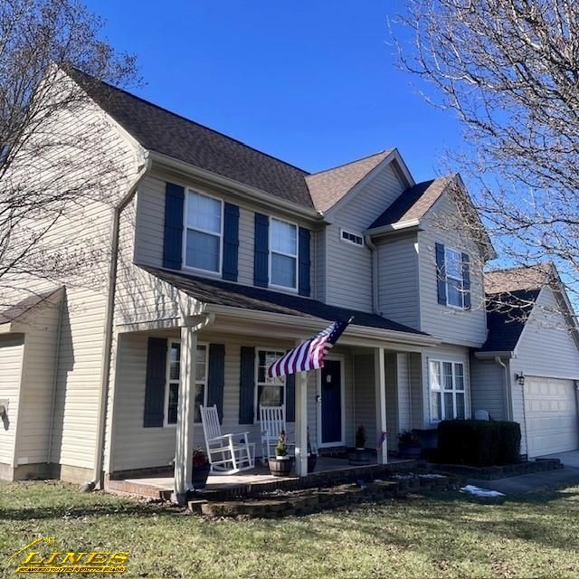 Two-story beige house with black roof, blue shutters, front porch with chairs, American flag, and a clear blue sky.