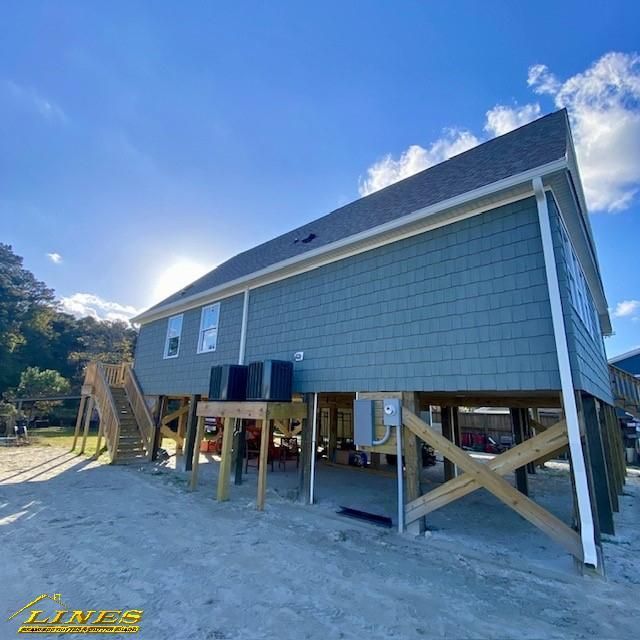Blue shingle house on stilts against a blue sky, with wooden stairs, and air conditioning units.