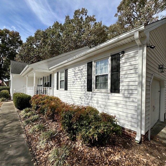 White house with black shutters, bushes, and a sidewalk, under a blue sky.