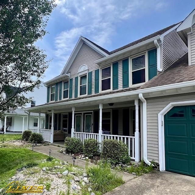 Two-story house with green shutters, garage door, and porch. Gray siding, green grass and shrubs. Sunny day.