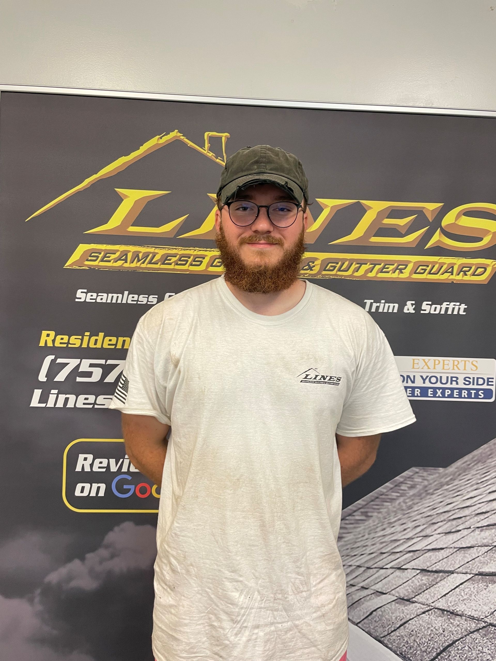 A man with a beard and glasses stands in front of a sign for a roofing company.