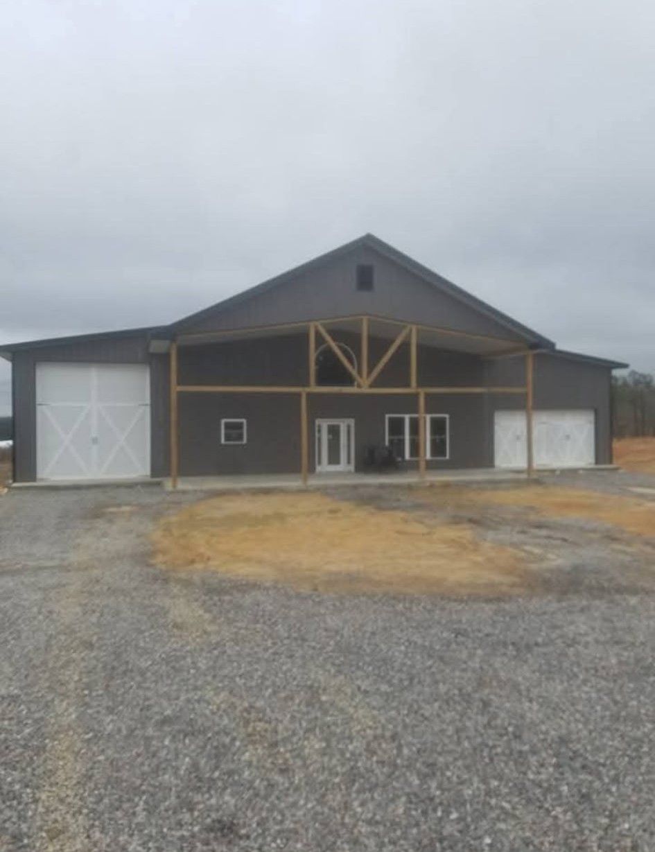 Gray barn-style building with white doors and a wooden frame. Set in a gravel lot under a cloudy sky.