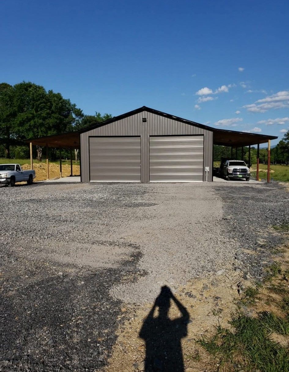 Gray metal garage with two bays, a lean-to, gravel driveway, and a blue sky.