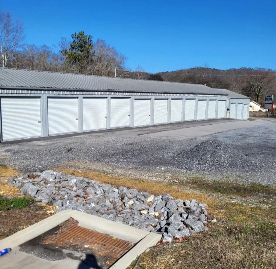 Storage units with white doors, gray roof, set in a gravel lot, with trees and hills in the background.