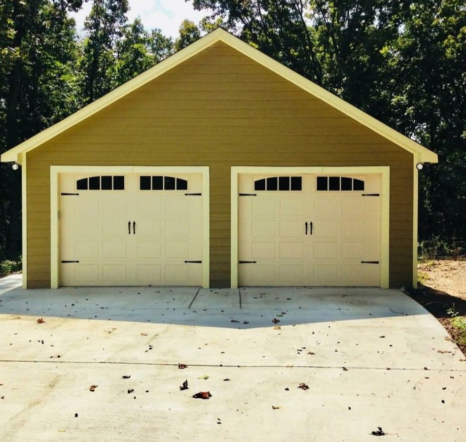 Two-car garage with light tan doors, olive siding, and concrete driveway, surrounded by trees.