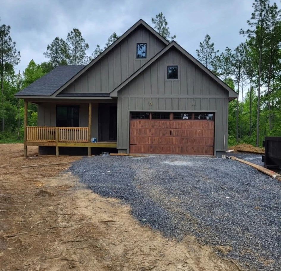 New house under construction; gray siding, brown garage door, small porch, gravel driveway, trees.