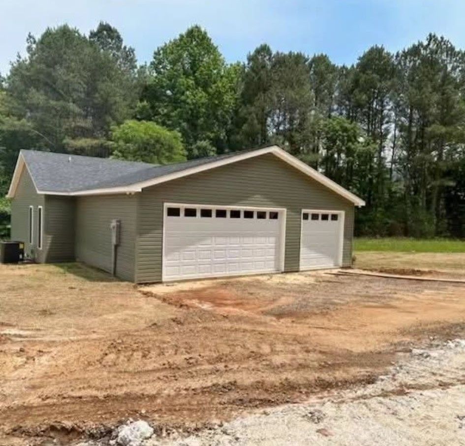 Two-car green garage with white doors, a dirt lot, and trees in the background.