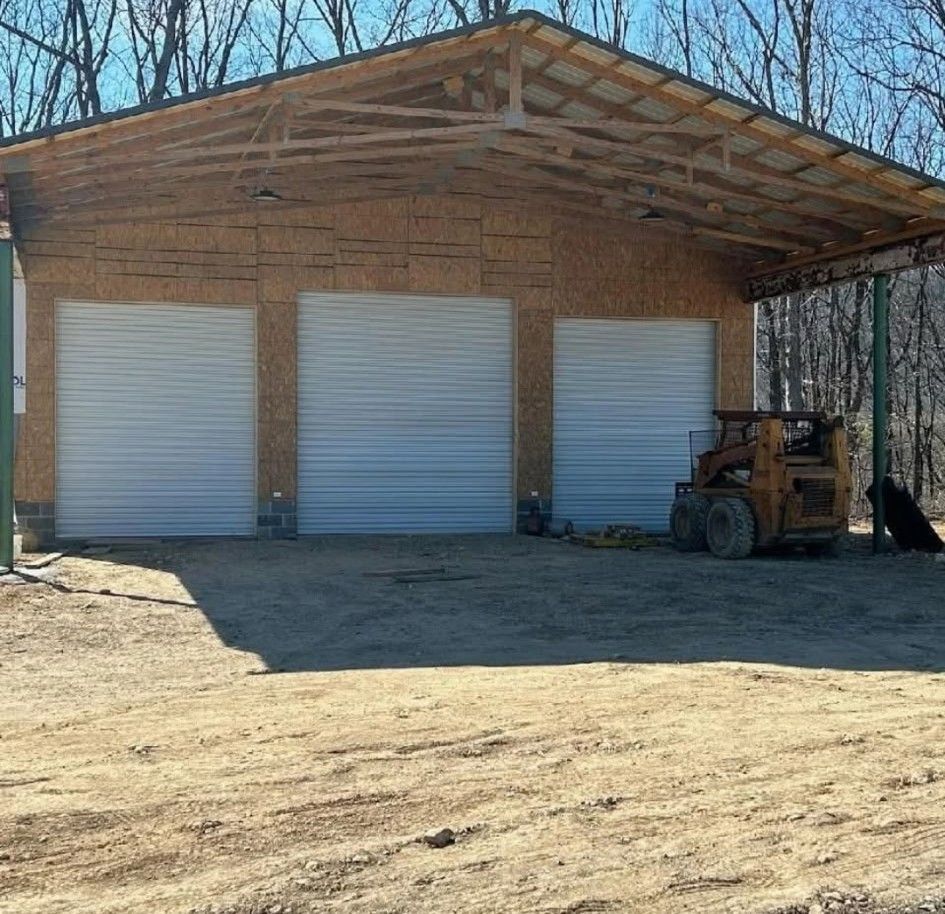 Three-bay garage with roll-up doors, wooden frame, metal roof, dirt driveway, small tractor, and woods in the background.
