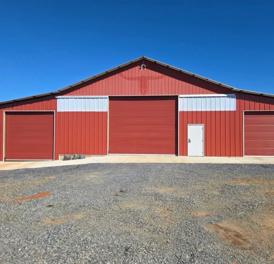 Red metal barn with three large doors, a small white door, and a gravel driveway under a blue sky.