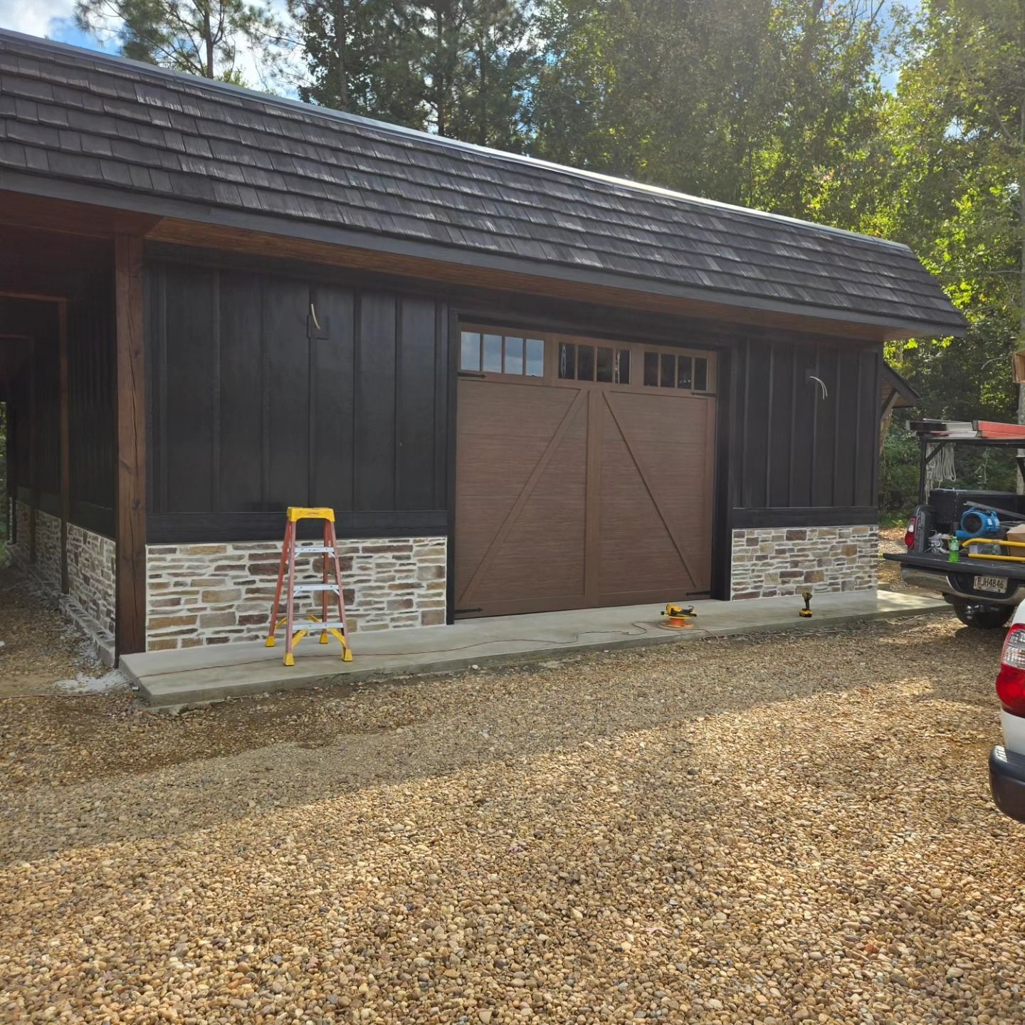 Dark wood garage with stone base, brown door, cedar shake roof, and gravel driveway.