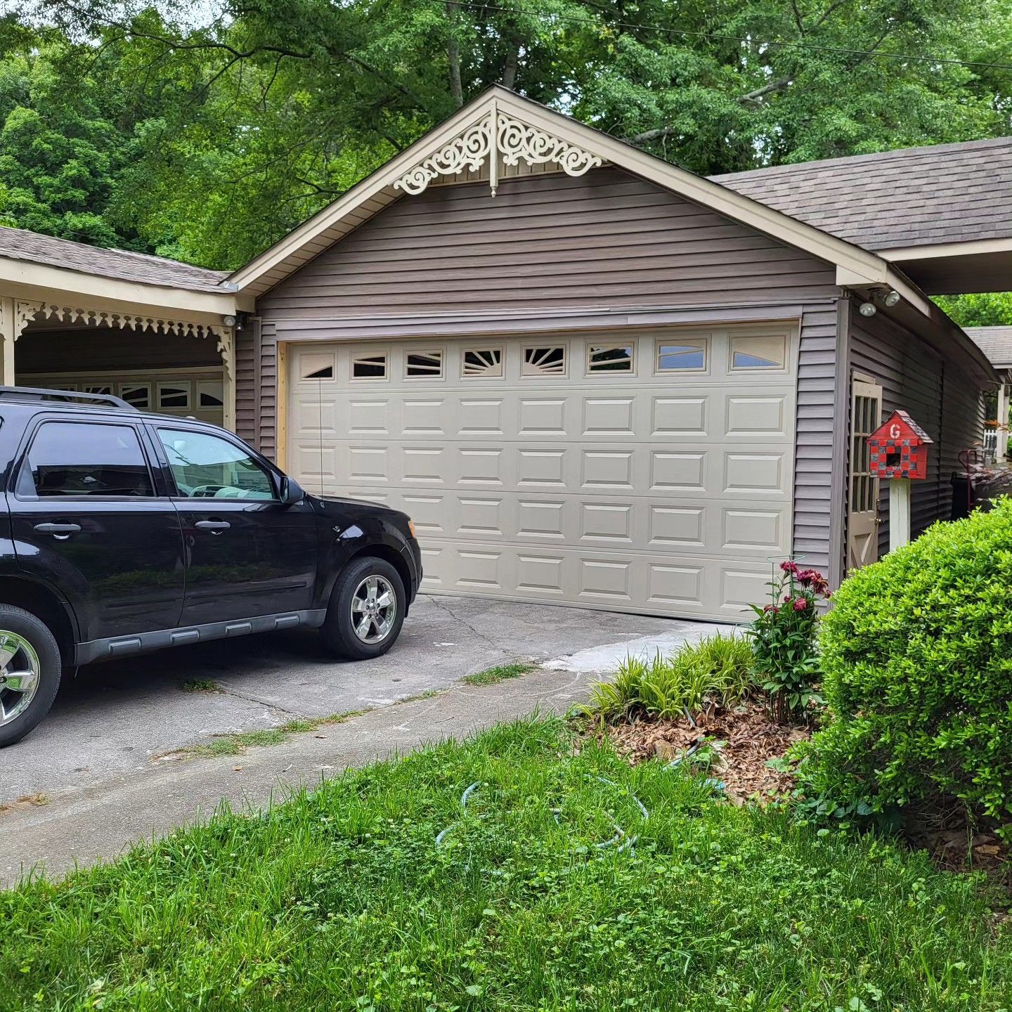 Black SUV parked in front of a beige garage with a brown roof. Green bushes and trees surround the driveway.