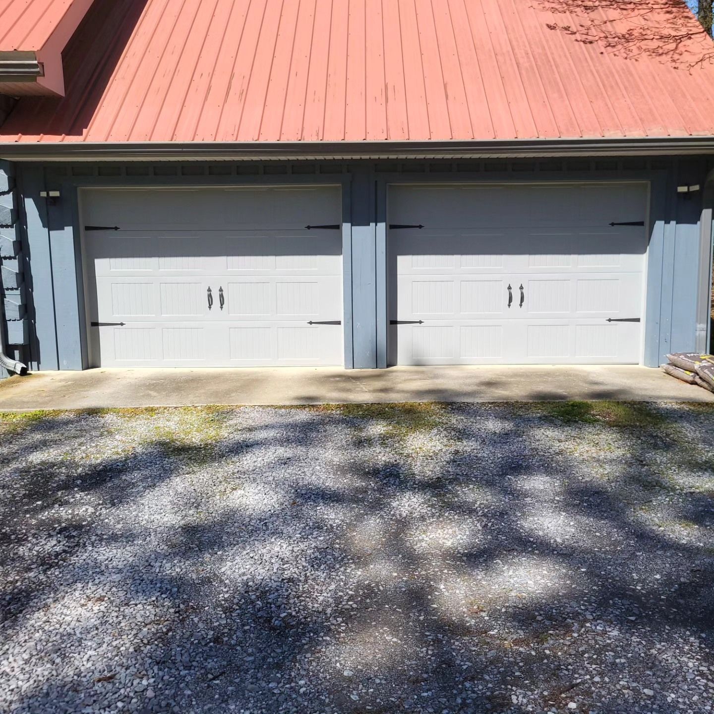 Two white garage doors on a building with a red roof and blue siding, set in a gravel driveway.