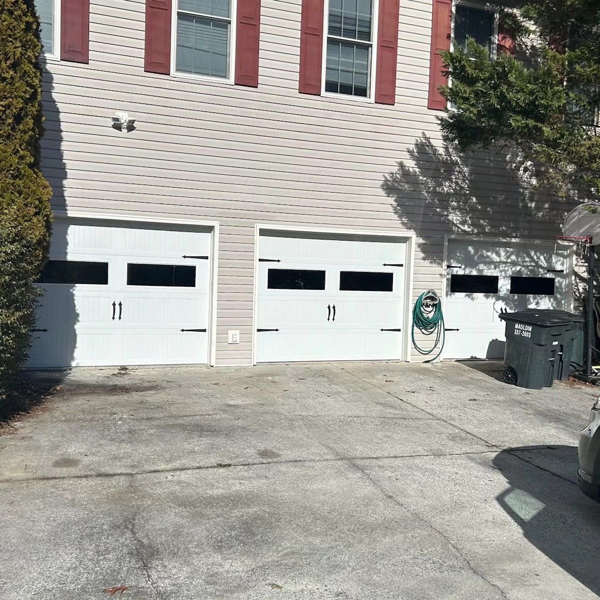 Three white garage doors on a house with light siding and red shutters. Driveway in front.