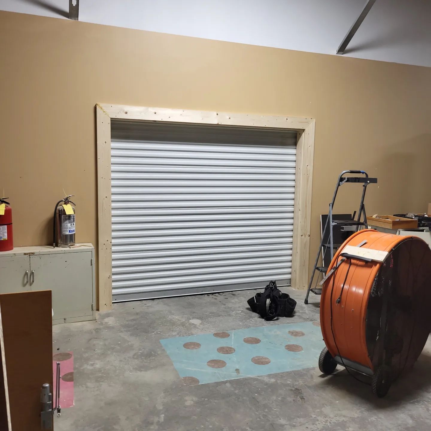 Interior view of a garage with closed metal roll-up door framed in wood.  A large orange fan sits nearby.
