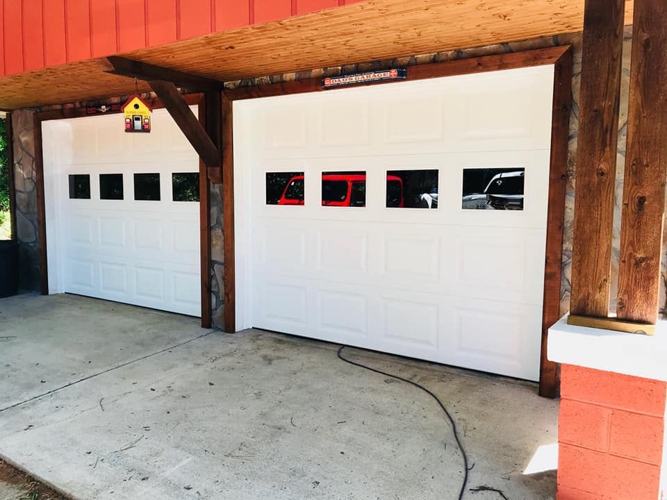 Two white garage doors with windows, under a wooden roof, set in a concrete area.