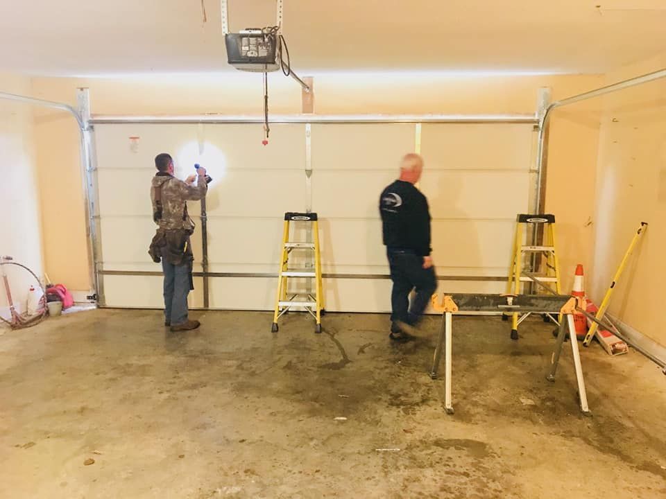 Two men installing a garage door in a garage; ladders and tools are present.
