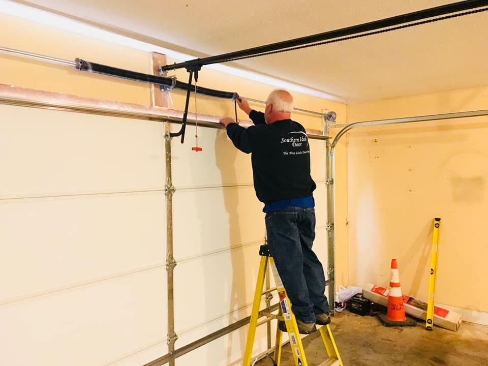 Man on a ladder repairing a garage door spring system in a garage.