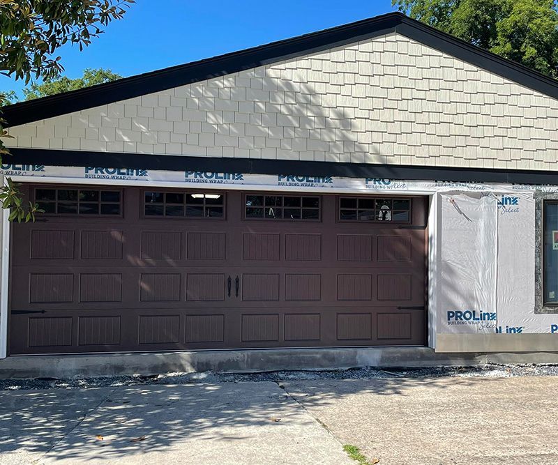 Brown garage door with decorative windows; tan shingle siding; construction in progress.