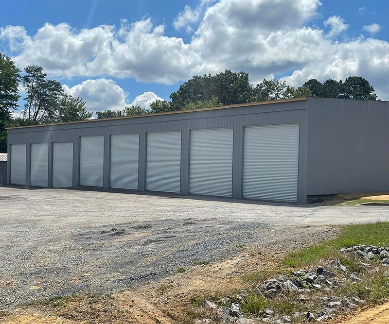 Storage units building with gray exterior and white roll-up doors, gravel lot, and cloudy sky.