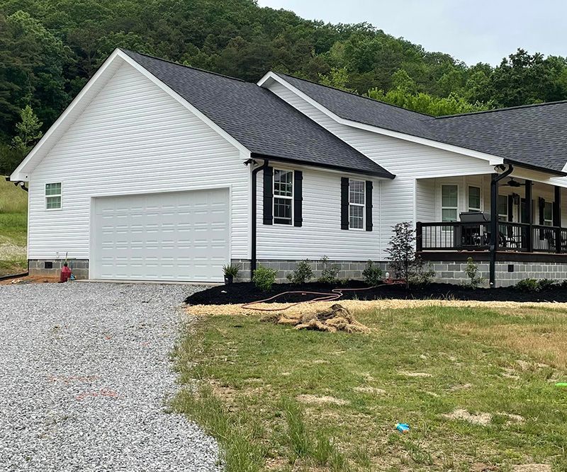 White house with black trim, garage, porch, and gravel driveway, set against a hillside.