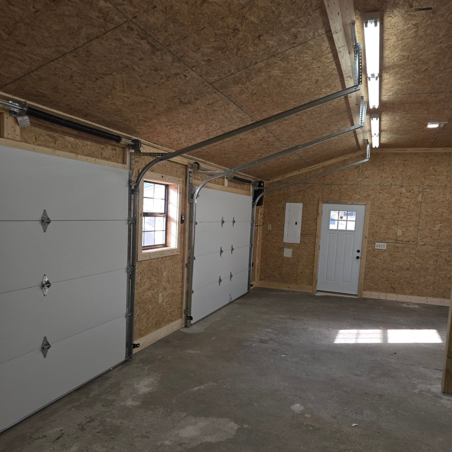 Inside of a new garage, two white garage doors, a small window, a white door, and concrete floor.