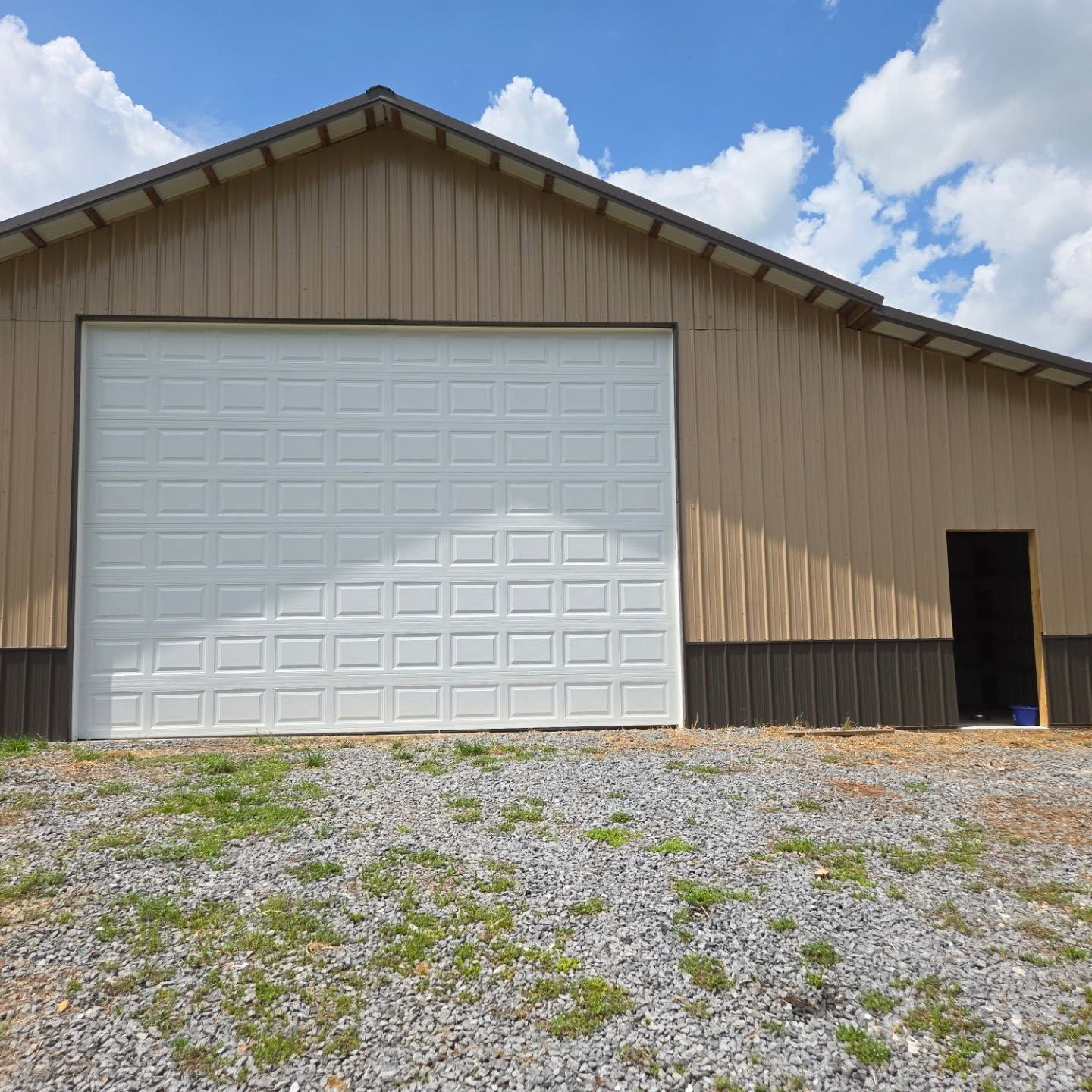 Tan and brown metal building with a large white garage door and a small open doorway on a gravel driveway.
