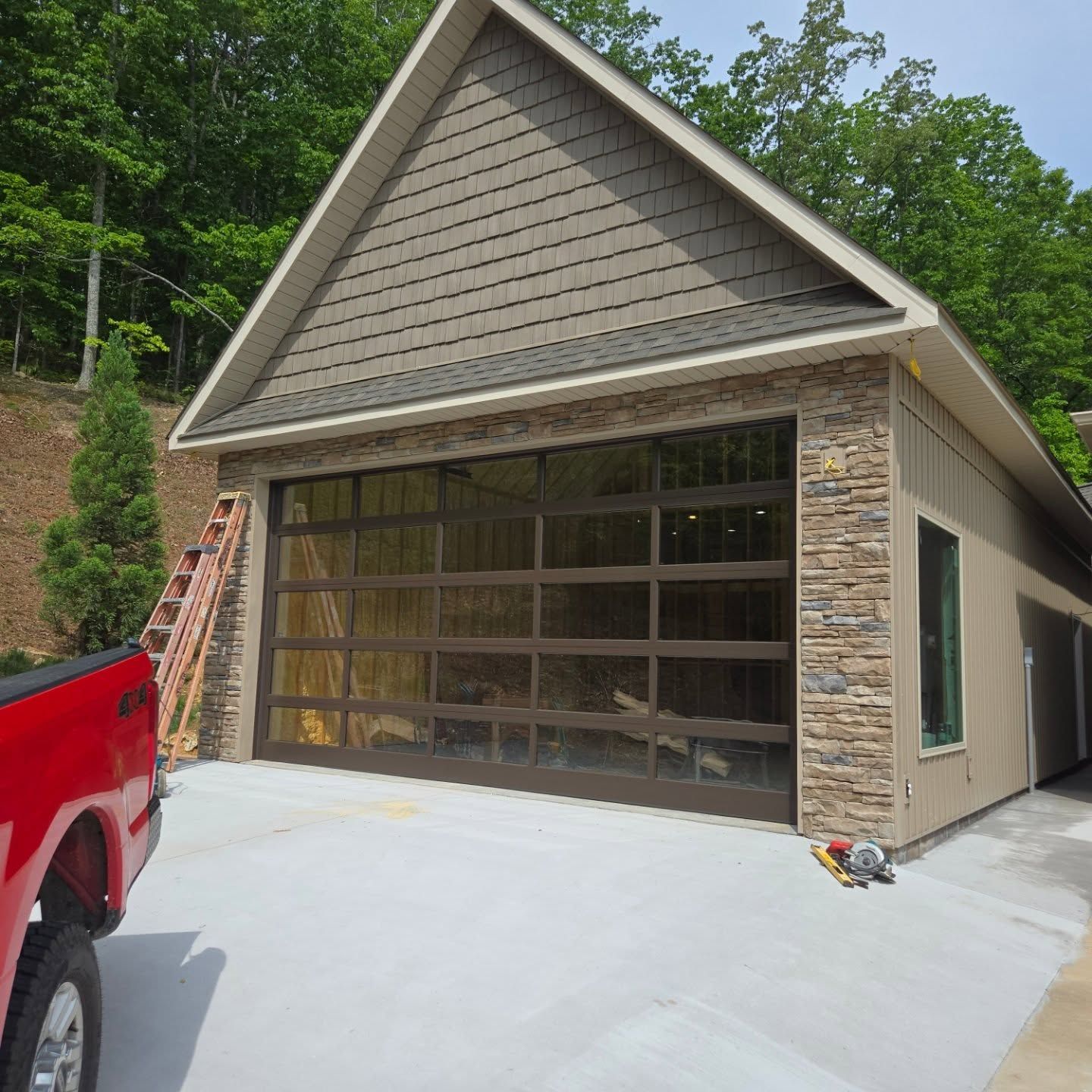 Garage with glass door and brown trim, attached to a beige building. Concrete driveway, red truck, and ladder present.