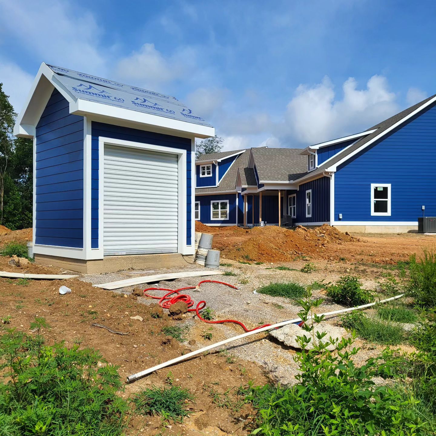 Blue shed with roll-up door next to a blue house under construction; sunny day, dirt foreground.