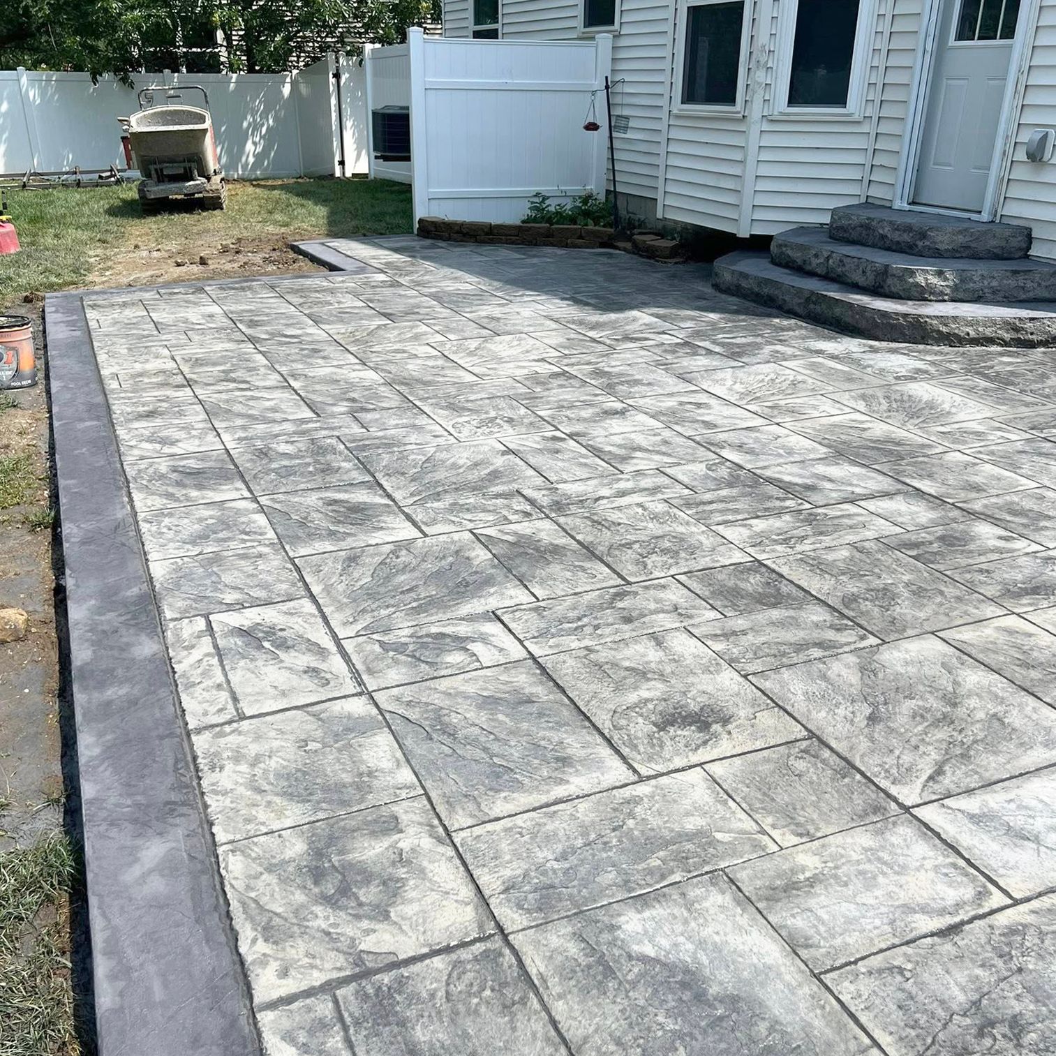 Gray paver patio with dark gray border, next to a white house, outdoors.
