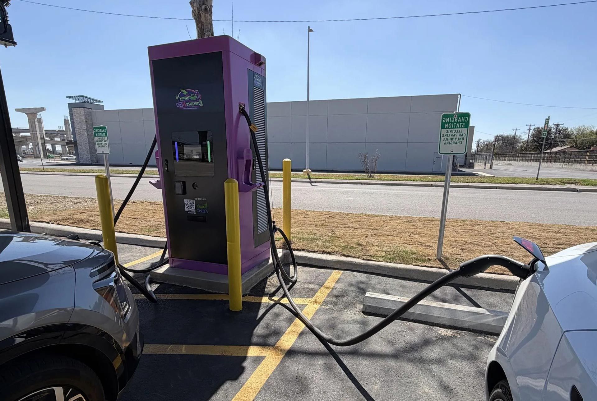 Electric vehicle charging stations lined along a curb under trees, with cables and control panels.