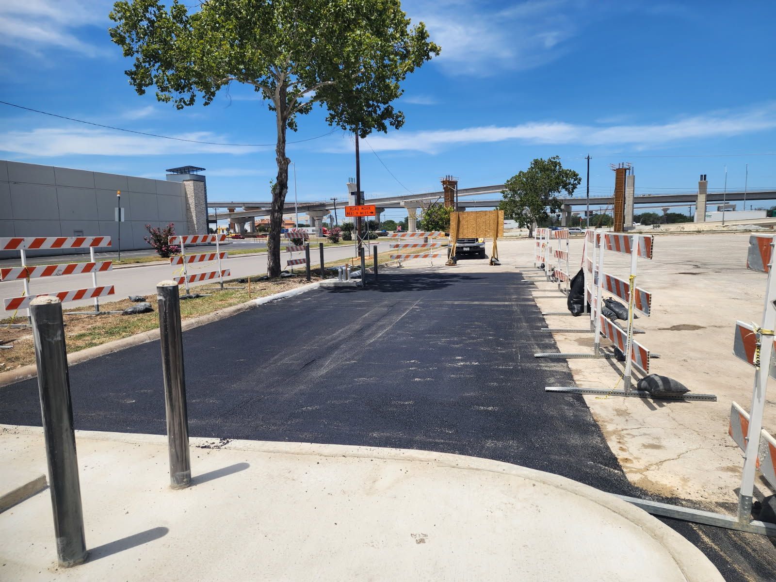 New pavement at a construction site with trees, barriers, and equipment under a clear blue sky