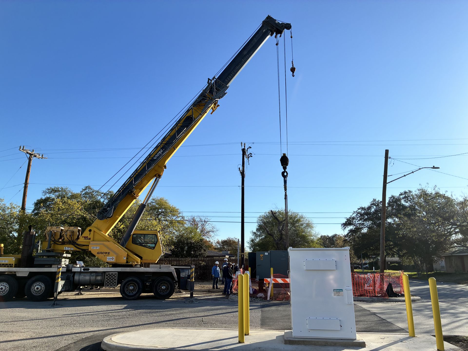 Yellow crane lifting hook at an outdoor work site beside a white utility box and utility poles under a blue sky