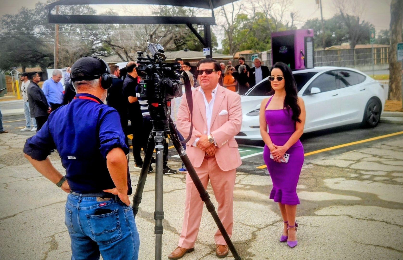 Outdoor interview setup with two people posing beside a camera crew and parked car in a wet lot.