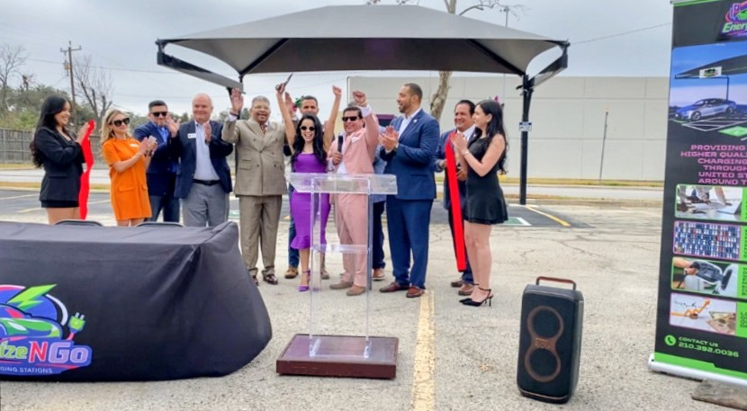 Group of people at an outdoor podium event under a canopy, with speakers and promotional banners on either side.