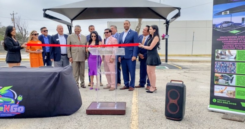Ribbon-cutting ceremony with people on a stage, red ribbon, microphone stand, and speakers outdoors