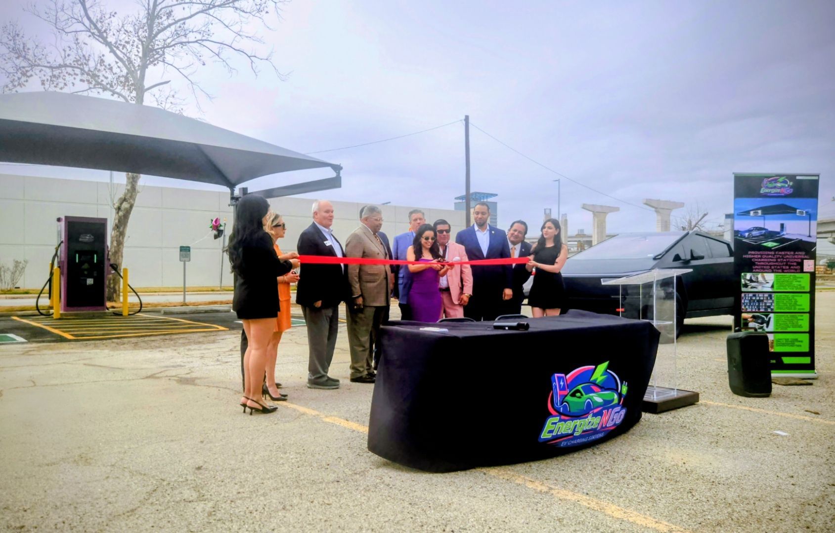 Ribbon-cutting ceremony outdoors with a group behind a red ribbon and a branded table.